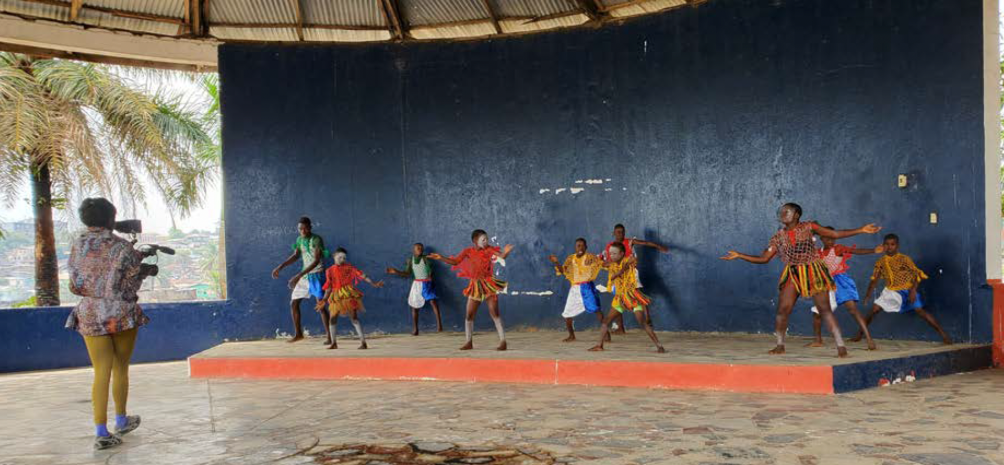 A woman holding a camer and filming as a group of children dance
