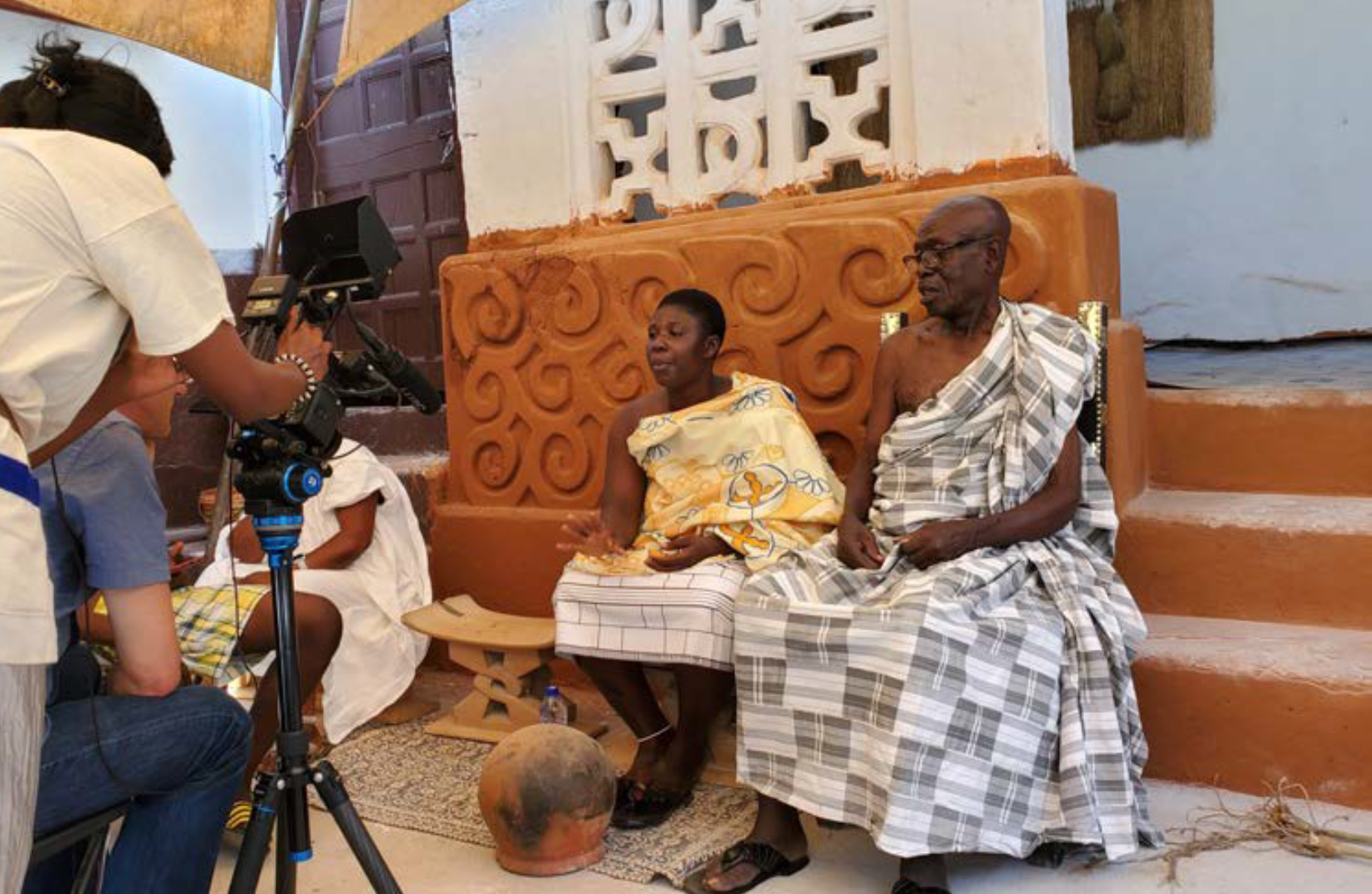 A woman and a man in traditional Ghanaian dress are seated on wooden stools and speak into a camera