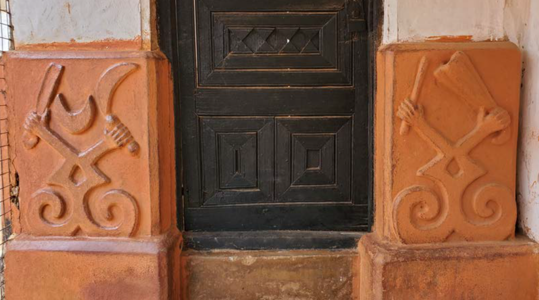 View of a building showing carved wooden door and clay walls, the upper parts white and the lower parts red, with raised geometric designs