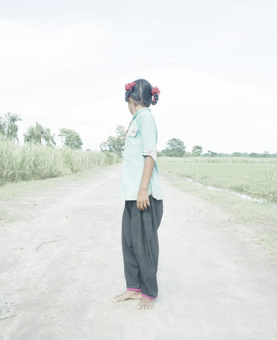 Straight-on view of a girl in braids and a blue shirt standing on a dirt rode and looking over her shoulder, away from the camera