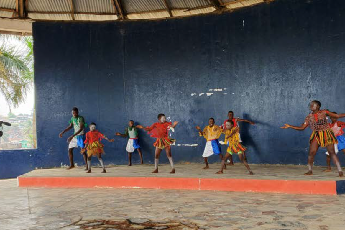 A woman holding a camer and filming as a group of children dance