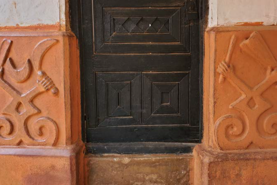 View of a building showing carved wooden door and clay walls, the upper parts white and the lower parts red, with raised geometric designs