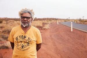 Straight-on view of an Aboriginal man in a yellow shirt standing in a desert along the roadside