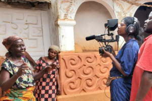 A woman holds a camera and films another woman speaking in front of an earthen building