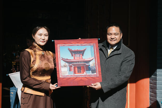 A man in a grey coat and a woman in a silk dress hold a photograph of a building while smiling.