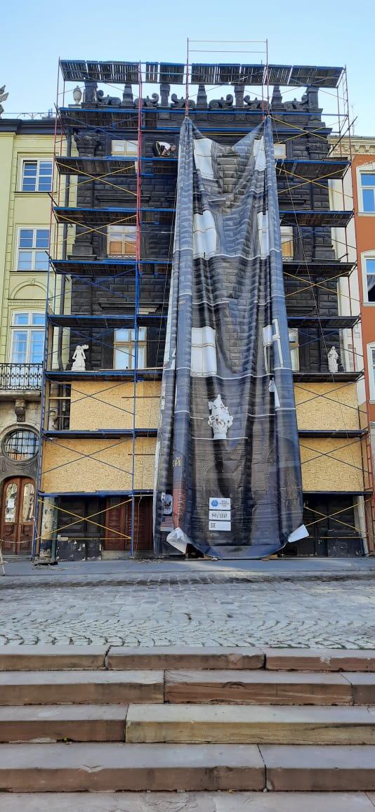 Tall black historic building with scaffolding over it. A tarp is being lifted over the scaffolding to cover it.