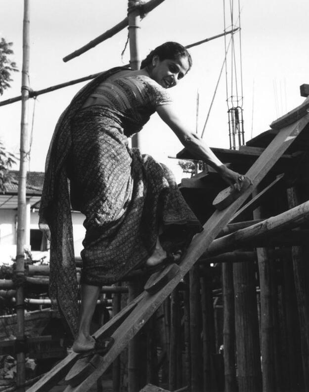 Black-and-white photo of young woman in sari climing a wooden ladder.